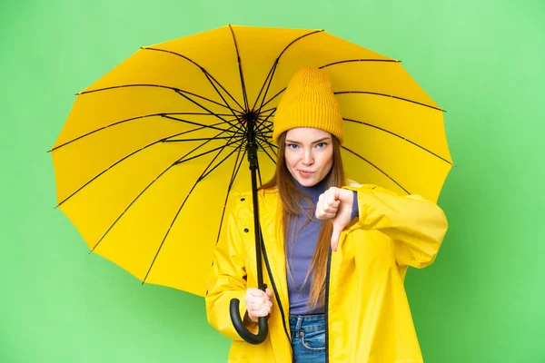 年轻漂亮的女人 穿着防雨外套 带着雨伞 罩在孤立的彩色关键背景上 带着负面表情垂下大拇指 — 图库照片