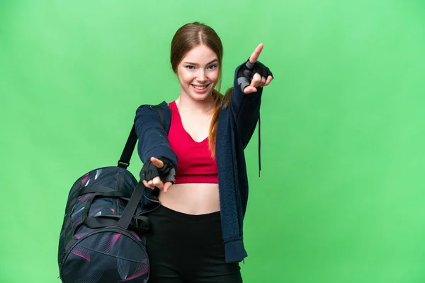 Young sport woman with sport bag over isolated chroma key background points finger at you while smiling