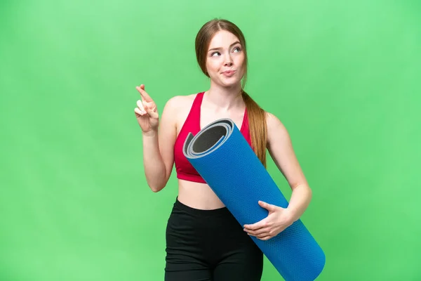 Young sport woman going to yoga classes while holding a mat over isolated chroma key background with fingers crossing and wishing the best