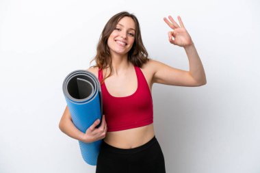 Young sport caucasian woman going to yoga classes while holding a mat isolated on white background showing ok sign with fingers