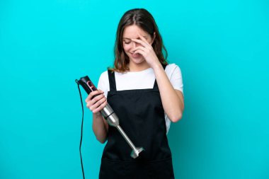 Beyaz tenli genç aşçı kadın el blenderi kullanıyor. Mavi arka planda izole edilmiş gülüyor.