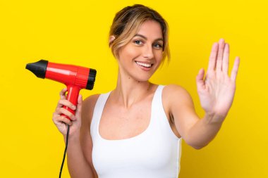 Young Uruguayan woman holding a hairdryer isolate don yellow background saluting with hand with happy expression