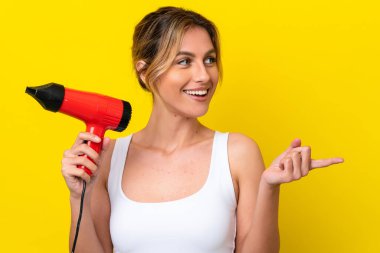 Young Uruguayan woman holding a hairdryer isolate don yellow background pointing to the side to present a product