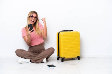 Uruguayan traveler woman sitting on the floor isolated on white background with phone in victory position
