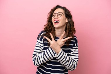 Pembe arka planda izole edilmiş genç beyaz kadın gülümsüyor ve zafer işareti gösteriyor.