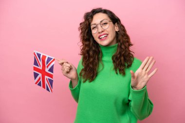 Young caucasian woman holding English flag isolated on pink background saluting with hand with happy expression