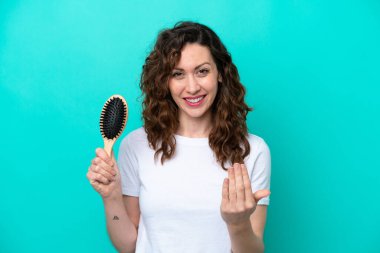 Young caucasian woman holding hairbrush isolated on blue background inviting to come with hand. Happy that you came