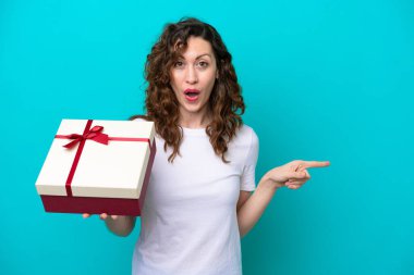 Young caucasian woman holding a gift isolated on blue background surprised and pointing side