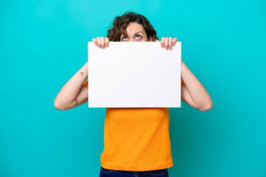 Kafkasyalı genç bir kadın mavi arka planda izole edilmiş boş bir levha tutuyor ve arkasında saklanıyor.