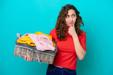 Young caucasian woman holding a clothes basket isolated on blue background having doubts and thinking