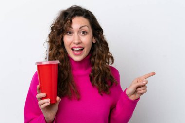 Young caucasian woman drinking soda isolated on white background surprised and pointing finger to the side