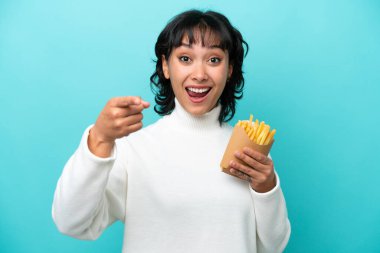 Young Argentinian woman holding fried chips isolated on blue background surprised and pointing front