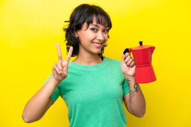 Young Argentinian woman holding coffee pot isolated on yellow background smiling and showing victory sign