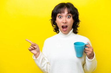 Young Argentinian woman holding cup of coffee isolated on yellow background surprised and pointing side