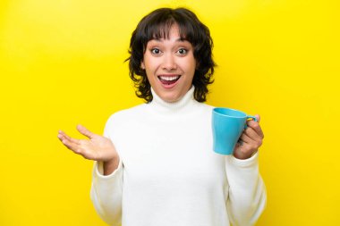Young Argentinian woman holding cup of coffee isolated on yellow background with shocked facial expression