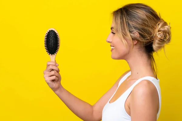 Young Uruguayan woman with hair comb isolated on yellow background with happy expression