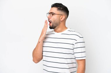 Genç Arap yakışıklı adam beyaz arka planda izole edilmiş esniyor ve ağzı açık bir şekilde elleriyle ağzını kapatıyor.