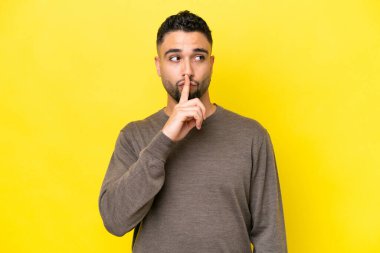 Sarı arka planda izole edilmiş genç Arap yakışıklı bir adam sessizlik işareti gösteriyor. Parmağını ağzına sokuyor.