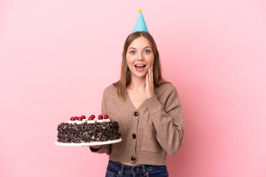 Young Lithuanian woman holding birthday cake isolated on pink background with surprise and shocked facial expression
