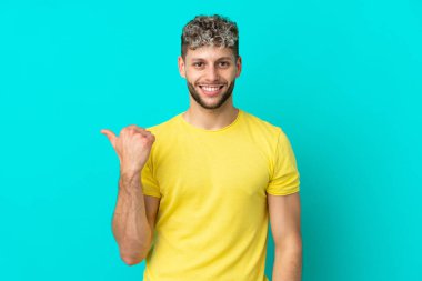 Mavi arka planda izole edilmiş yakışıklı beyaz adam bir ürünü sunmak için kenarı işaret ediyor.