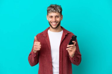 Young handsome caucasian man isolated on blue background using mobile phone while doing thumbs up