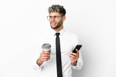 Beyaz arka planda izole edilmiş beyaz bir iş adamı elinde kahve ve bir şeyler düşünürken cep telefonu tutuyor.