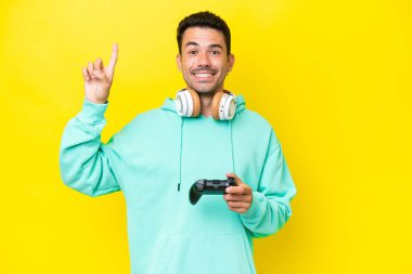 Young handsome man playing with a video game controller over isolated wall pointing up a great idea