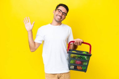 Young handsome man holding a shopping basket full of food over isolated yellow background saluting with hand with happy expression