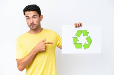 İzole edilmiş beyaz arka plan üzerinde genç yakışıklı bir adam elinde geri dönüşüm simgesiyle bir pankart tutuyor ve onu işaret ediyor.