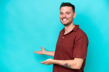 Beyaz tenli yakışıklı adam mavi arka planda izole edilmiş. Davet etmek için ellerini yana uzatıyor.