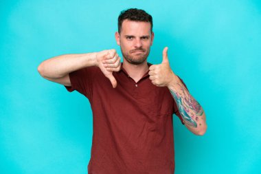 Beyaz tenli yakışıklı adam mavi arka planda izole edilmiş, iyiye-kötüye işaret yapıyor. Evet ya da hayır arasında kararsız