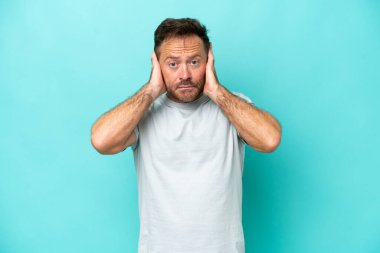 Mavi arka planda izole edilmiş orta yaşlı beyaz adam hayal kırıklığına uğramış ve kulaklarını kapatmış.