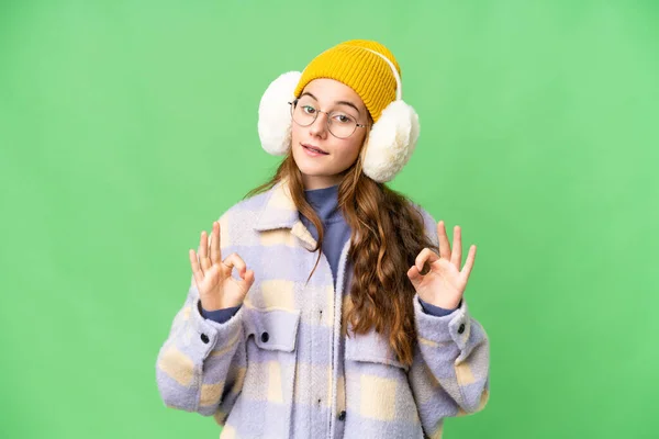 Teenager girl wearing winter muffs over isolated chroma key background showing an ok sign with fingers