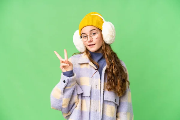 Teenager girl wearing winter muffs over isolated chroma key background smiling and showing victory sign