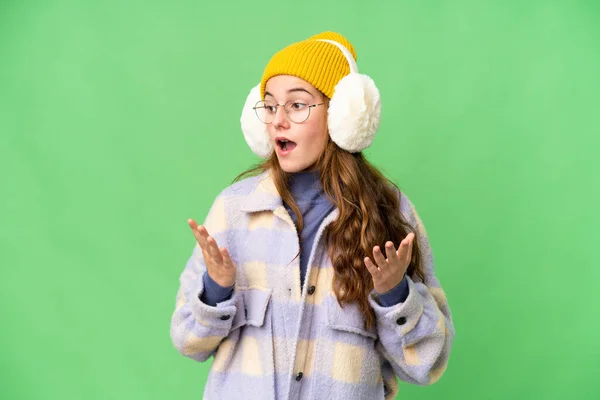 Teenager girl wearing winter muffs over isolated chroma key background with surprise facial expression
