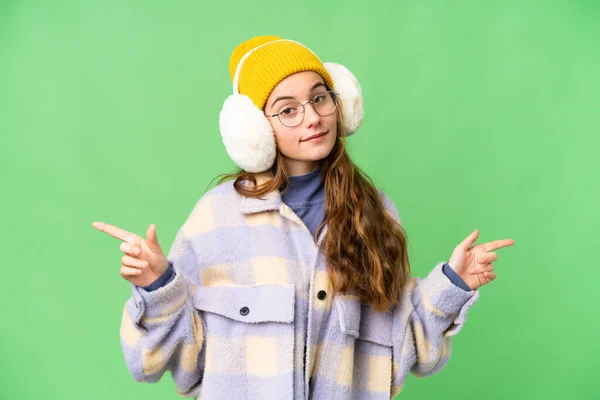 Teenager girl wearing winter muffs over isolated chroma key background pointing finger to the laterals and happy