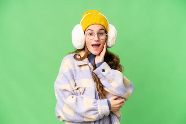 Teenager girl wearing winter muffs over isolated chroma key background surprised and shocked while looking right