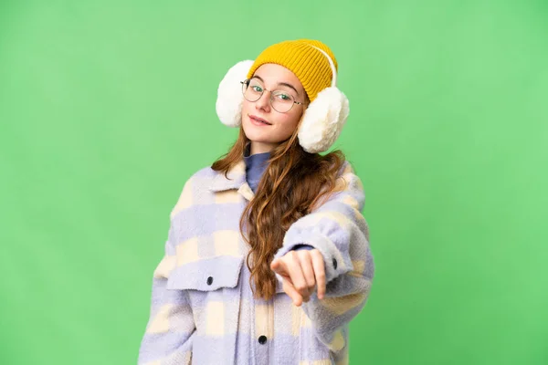 Teenager girl wearing winter muffs over isolated chroma key background points finger at you with a confident expression