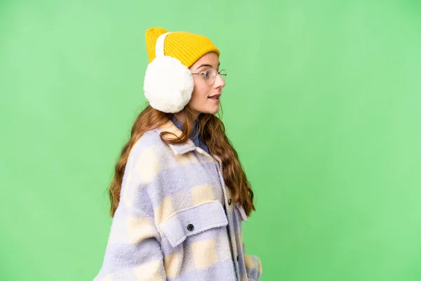 Teenager girl wearing winter muffs over isolated chroma key background laughing in lateral position