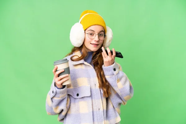 Teenager girl wearing winter muffs over isolated chroma key background holding coffee to take away and a mobile