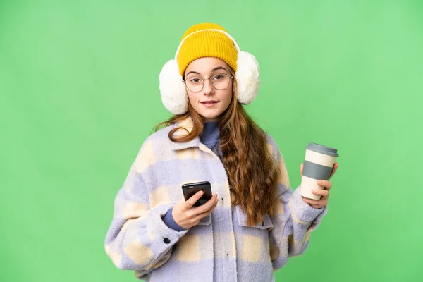 Teenager girl wearing winter muffs over isolated chroma key background holding coffee to take away and a mobile