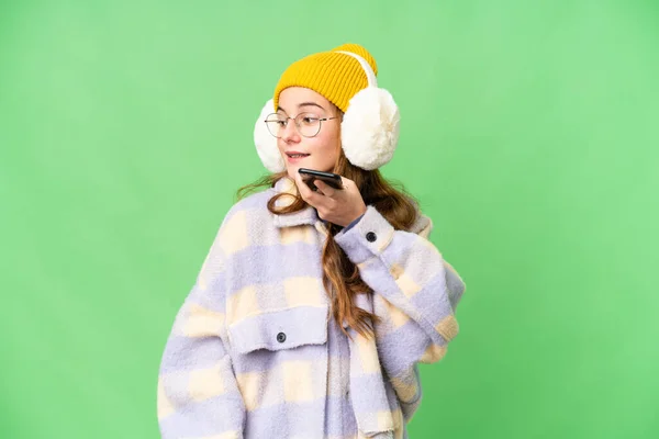 Teenager girl wearing winter muffs over isolated chroma key background keeping a conversation with the mobile phone with someone