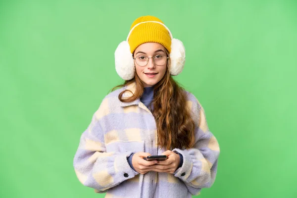 Teenager girl wearing winter muffs over isolated chroma key background sending a message with the mobile