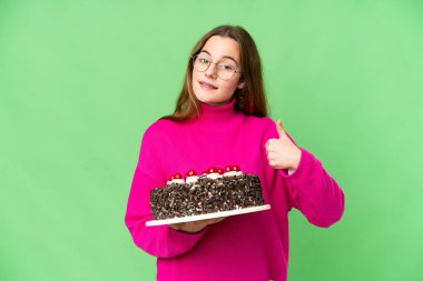 Teenager girl holding birthday cake over isolated chroma key background giving a thumbs up gesture