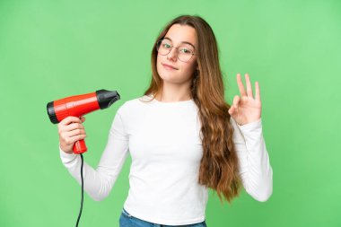 Teenager girl holding a hairdryer over isolated chroma key background showing ok sign with fingers
