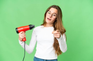 Teenager girl holding a hairdryer over isolated chroma key background surprised and pointing front