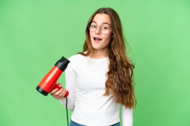Teenager girl holding a hairdryer over isolated chroma key background with surprise and shocked facial expression