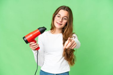 Teenager girl holding a hairdryer over isolated chroma key background points finger at you with a confident expression