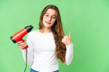 Teenager girl holding a hairdryer over isolated chroma key background pointing to the side to present a product