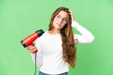 Teenager girl holding a hairdryer over isolated chroma key background having doubts and with confuse face expression